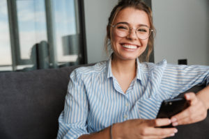 Image of happy young woman in eyeglasses smiling and using phone while sitting on settee
