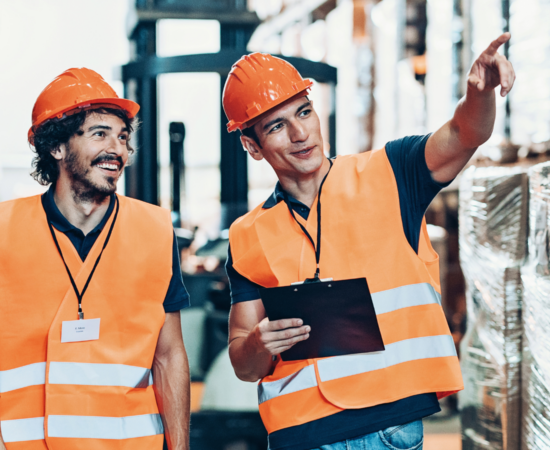 Man giving a safety talk to another man in a warehouse wearing high vis vests and hard hats
