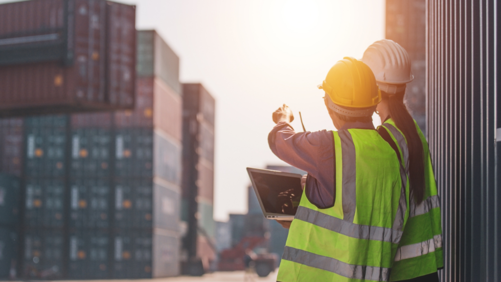 Two men stood on a port site with a clipboard pointing at cargo