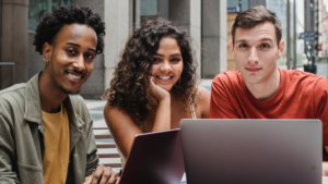 Three students facing forward and smiling whilst sat at laptops