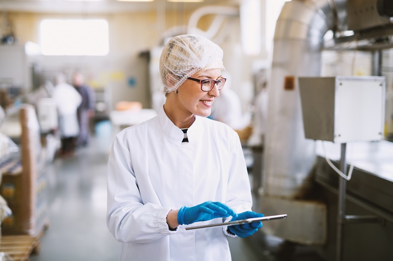 Smiley female worker in sterile clothes using tablet and checking how production line is working.