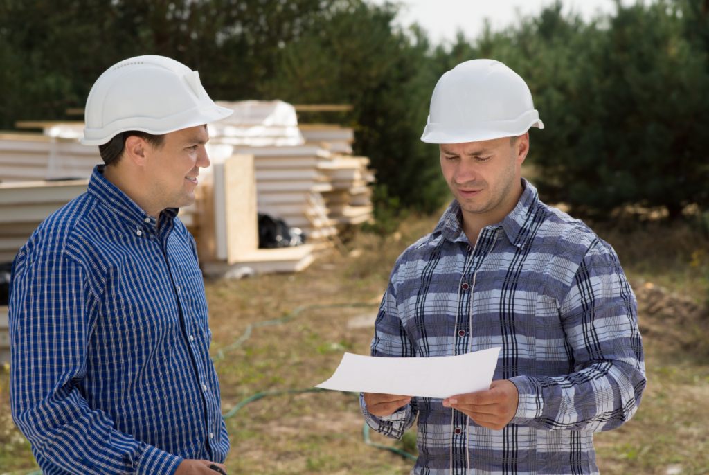 Two men in hard hats stood on a construction site reading blueprints