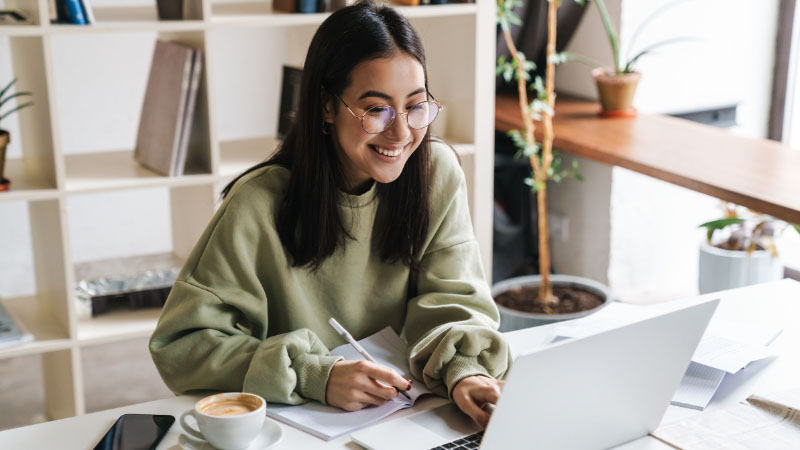 Smiling student sat at laptop taking notes